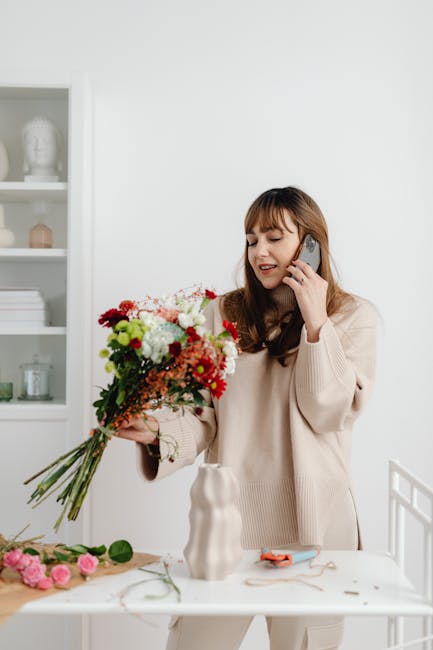 A woman with light brown hair wearing a beige sweater is holding a large, vibrant floral bouquet featuring white, red, and pink flowers along with green foliage. She is smiling and talking on a mobile