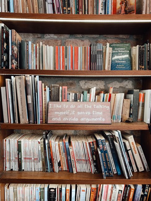 A wooden bookshelf filled with a variety of books arranged vertically and horizontally across multiple shelves. On the middle shelf, there is a pink and white sign with the humorous quote, 'I like to 