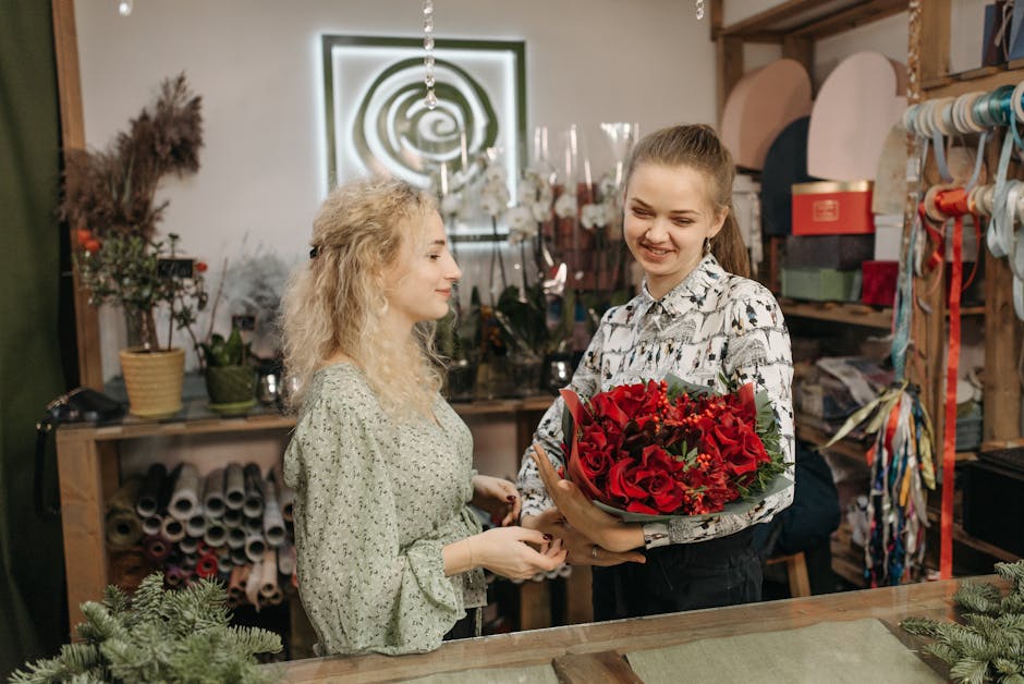 A young woman in a patterned blouse receives a vibrant bouquet of red roses, lilies, and greenery from a florist inside a floral shop. The bouquet is wrapped in green foliage and decorative paper. The