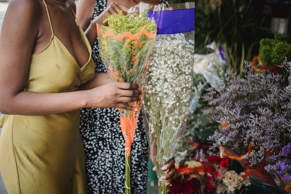 A person wearing a yellow spaghetti strap dress is holding a large bouquet of small green and white flowers wrapped in clear and orange floral paper, with a person in a black and white patterned outfi