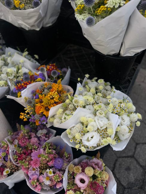 A selection of fresh flower bouquets displayed outdoors on a black crate surface, showcasing a variety of vibrant and pastel-colored flowers. The arrangements include white roses and chrysanthemums, p